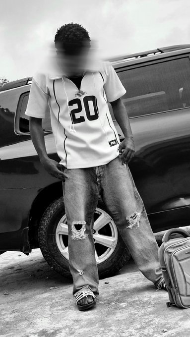 A black-and-white photograph of a person wearing a white sports jersey with the number 20 and ripped jeans, standing in front of an SUV with their face blurred.