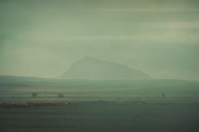 A solitary structure sits in a vast, hazy green landscape with a misty mountain rising in the background, evoking a quiet morning spent in nature. The image complements the caption "Good morning fam • Time Outside Time 1/1 350 XTZ" by capturing a serene outdoor scene that reflects a peaceful moment away from the busy world.