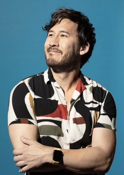 A portrait of a smiling man with dark hair and a beard, wearing a patterned short-sleeve shirt, standing against a solid blue background.