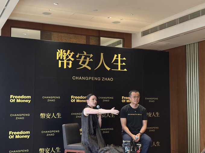A woman gesturing while speaking into a microphone alongside a seated man at a panel event with a backdrop featuring 'CHANGPENG ZHAO' and 'FREEDOM OF MONEY' in both English and Chinese characters.