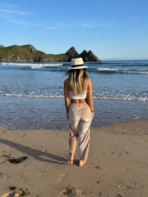 A woman with blonde hair, wearing a white top, beige pants, and a straw hat, stands on a sandy beach looking out at the ocean and rocky islands.