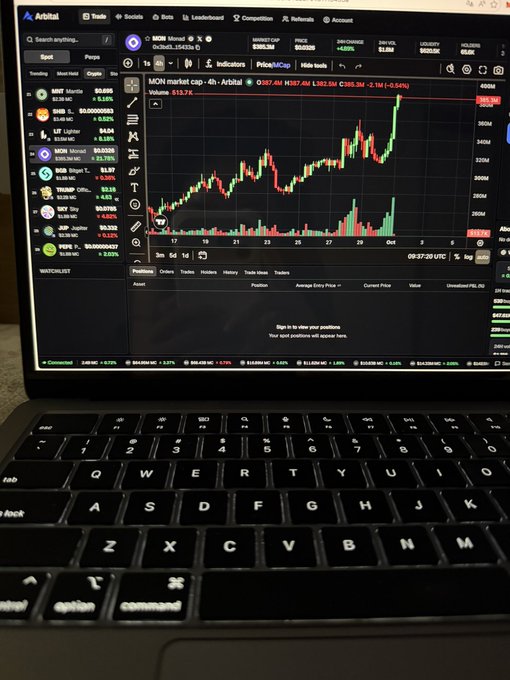 A photo of a MacBook screen showing the Arbitrum trading interface with the MOON memecoin chart displayed, alongside a watchlist of various cryptocurrencies.
