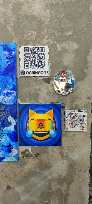 Street art on a concrete wall featuring a crying cat emoji sticker, a QR code labeled 'OGRINGO.75', a 'YOU ARE PART OF THIS CITY' badge, and blue painted tiles.