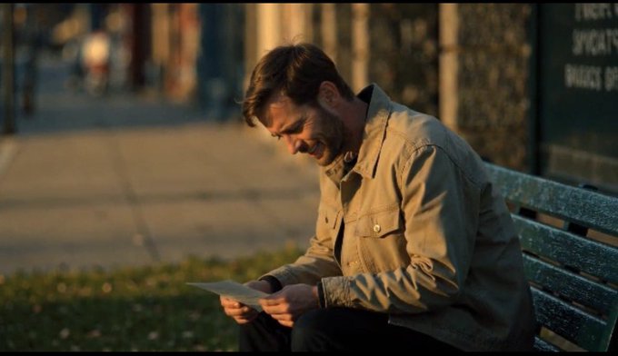 A man in a tan jacket sits on a green park bench during golden hour, smiling down at a piece of paper in his hands.
