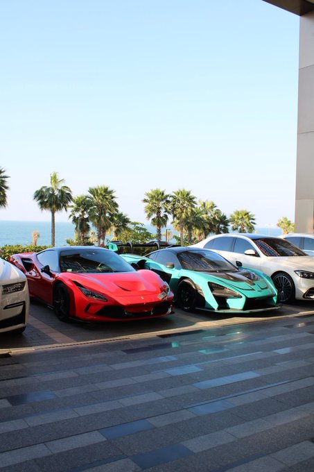 A red Ferrari and a teal McLaren are parked side by side on a tiled terrace overlooking the ocean, with palm trees lining the background. The luxury supercar display serves as a perfect visual "GM" (good morning) greeting, showcasing an impressive car meet in a scenic coastal setting.