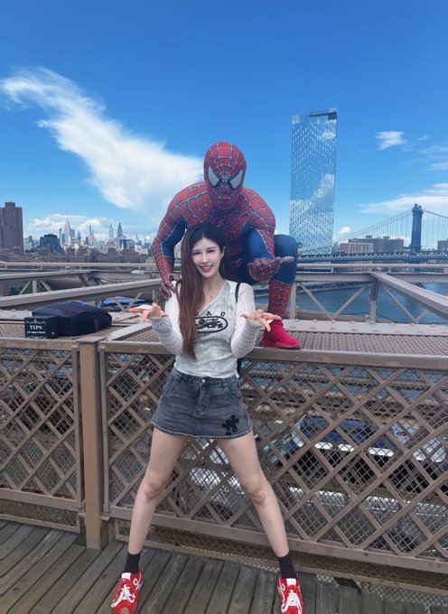 A young woman poses on the Brooklyn Bridge with someone dressed as Spider-Man behind her, with the Manhattan skyline visible in the background.