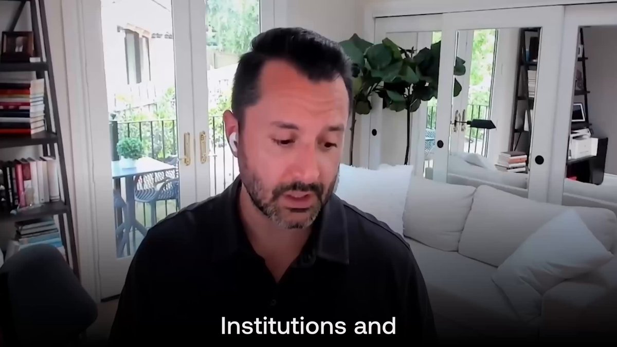 The image shows a man sitting in front of a couch with books scattered around him. The text "Institutions and" appears at the top, indicating that he might be discussing or talking about institutions. The man's facial expression suggests deep thought or contemplation as he looks down at his hands.