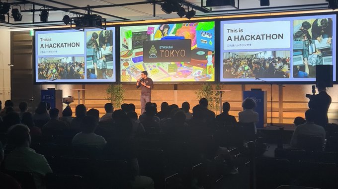 A presenter speaks on stage at a hackathon event, with large screens behind displaying "This is A HACKATHON" and "TOKYO".