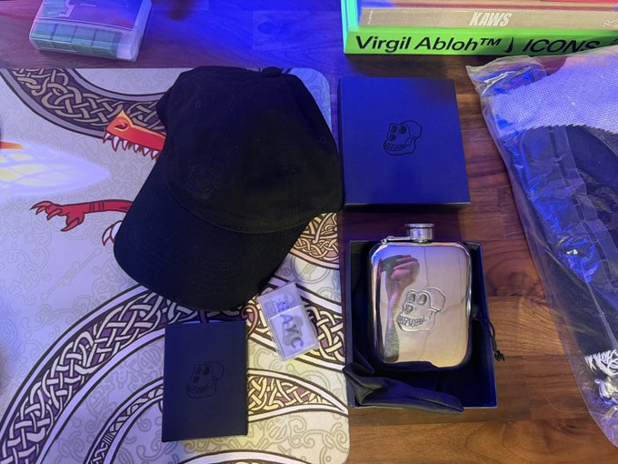 A collection of black-themed merchandise including a cap, a skull flask, and boxes, arranged on a table next to a Virgil Abloh 'ICONS' book and a playing mat.
