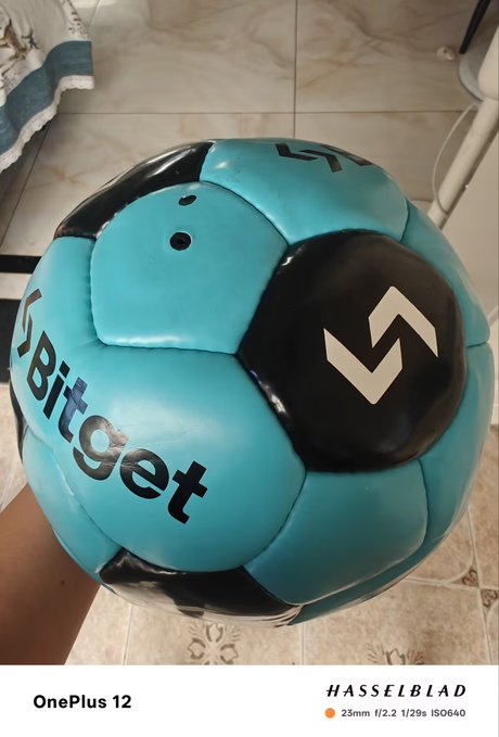 A close-up photo of a teal and black soccer ball with 'Bitget' branding and the Bitget logo, photographed with a OnePlus 12 (Hasselblad) camera on a tiled floor.