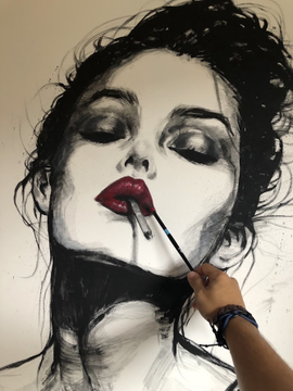 A black and white painting of a woman with closed eyes and bold red lips, with a hand holding a paintbrush actively working on the artwork.