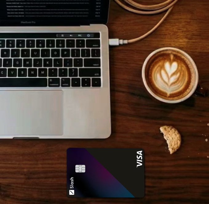 An overhead shot of a wooden desk with a MacBook Pro, a cup of coffee with latte art, a biscuit, and a Slash Visa debit card.