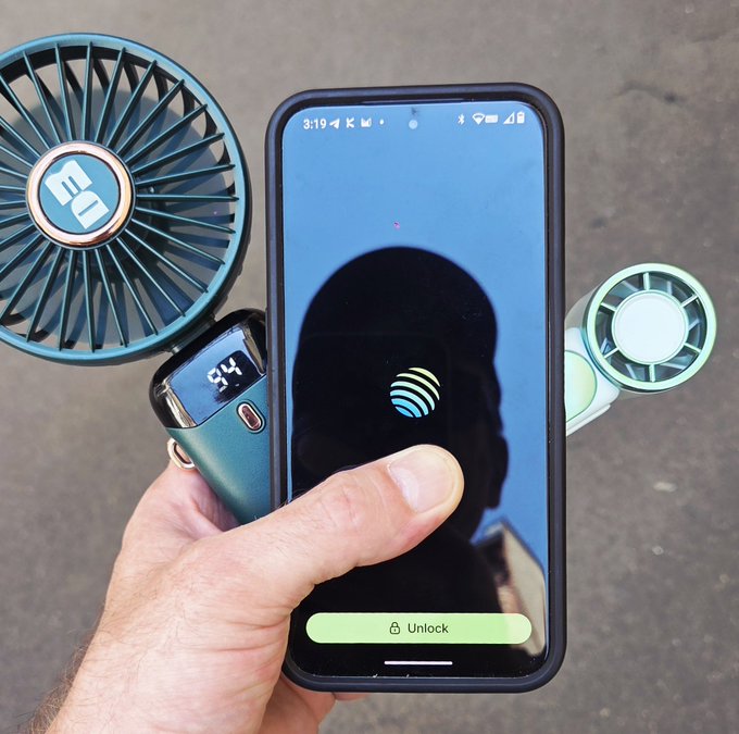 A hand holds a smartphone displaying an AT&T unlock screen with a fingerprint icon, alongside a portable fan showing "54°" and a "EO" branded fan, illustrating the post about using a Jupiter mobile fan for cooling in hot weather.