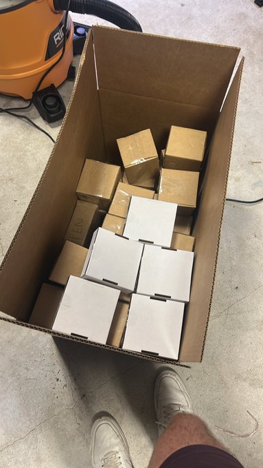 A photo looking down at an open cardboard box filled with smaller white and brown cardboard boxes, next to an orange RIDGID vacuum and a person's white sneakers.