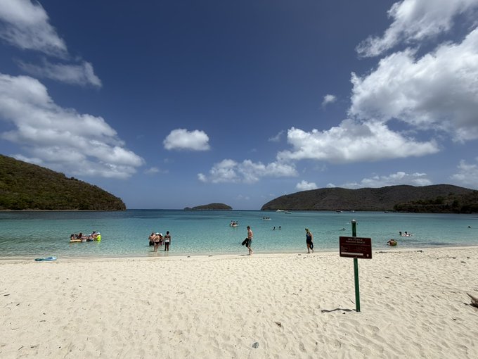 A pristine white-sand beach with turquoise waters and lush green hills frames swimmers enjoying the calm bay under a bright blue, cloud-dotted sky—an ideal scene for a relaxing weekend getaway.