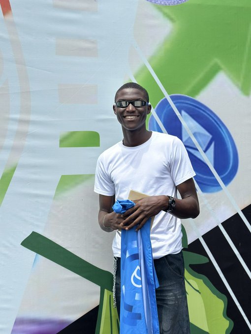 A smiling young man in a white t-shirt and dark glasses stands holding a blue flag/banner in front of a colorful geometric background.