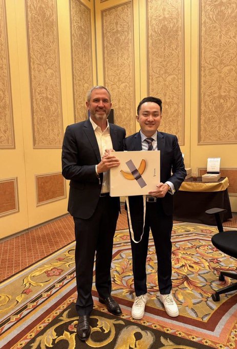 Two men in suits pose together holding a gift bag with a yellow banana graphic in an ornate room with patterned carpet.