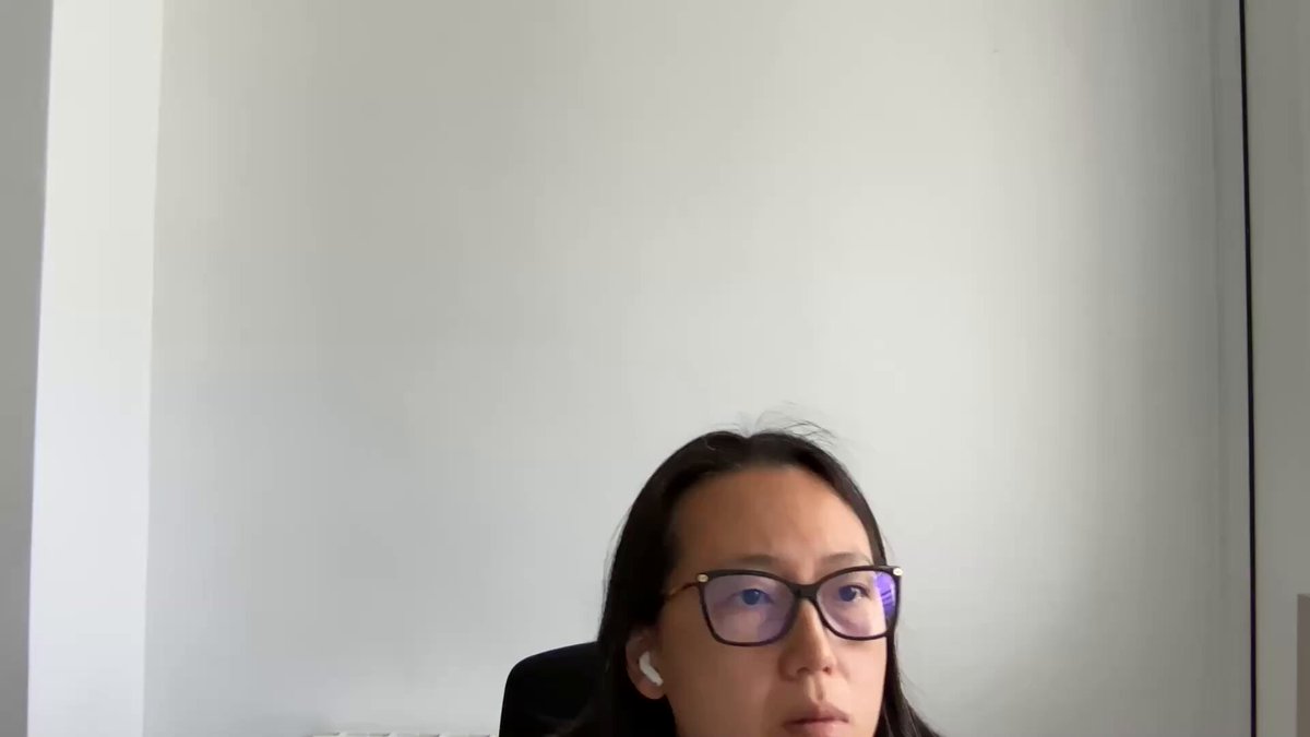 The image shows a woman wearing glasses sitting at a desk with her head tilted back as she looks up into the camera. The background of the photo is white, which contrasts with the black chair that the woman is seated on. There are no other people or objects in the scene, and the focus is solely on the woman's face and posture.