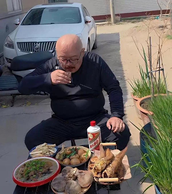 A bald man with glasses sitting outdoors drinking from a small glass, with a spread of dishes including fish, eggs, and greens on the table before him.