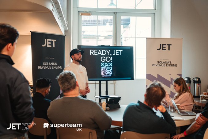 A man speaks to an audience at a 'JET' presentation, with banners reading 'JET Solana's Revenue Engine' and a screen showing 'READY, JET GO!' with a QR code for JetPitstop LDN and Superteam UK.