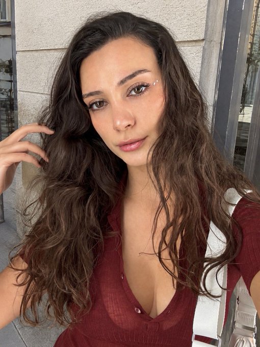 A young woman with long wavy brown hair poses for a selfie, wearing a deep red top with a low neckline and subtle eyebrow jewelry.