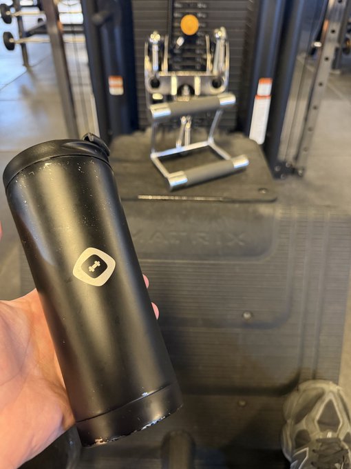 A hand holds a black insulated tumbler with a white logo in front of Matrix gym equipment, including a weight bench and rack, with a person's shoe visible at the bottom right.