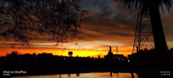 A stunning sunset photo shot on OnePlus by MSK on 10/24/23 at 5:46 PM, capturing silhouetted trees, power lines, and a water tower against a vibrant orange sky, reflecting the poster's passion for nature and photography taken in 2023.