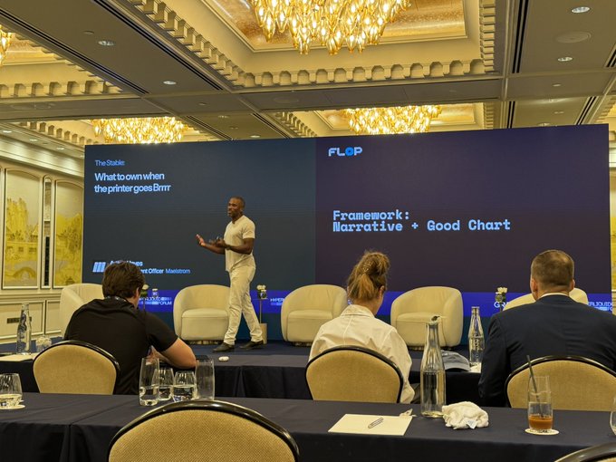 A presenter stands on stage at a conference, with slides behind him reading "The Stable: What to own when the printer goes Brrrr" and "FLOP Framework: Narrative + Good Chart", while panelists sit in chairs at the front.