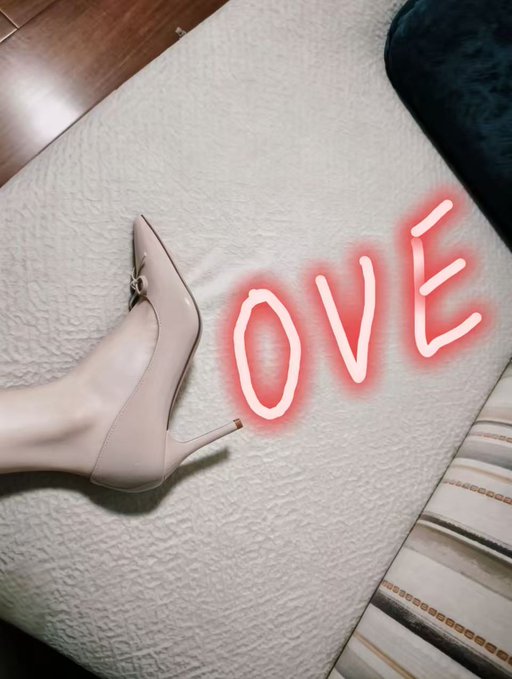 A first-person view of a foot wearing a nude pointed-toe stiletto heel with a gold horsebit detail, positioned on a beige textured mattress with a red neon 'OVE' sign glowing nearby.