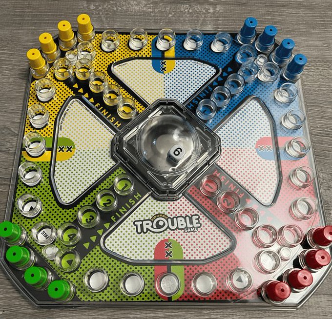 An overhead view of a Trouble board game featuring the iconic Pop-O-Matic dice roller in the center, surrounded by yellow, blue, green, and red colored pegs.