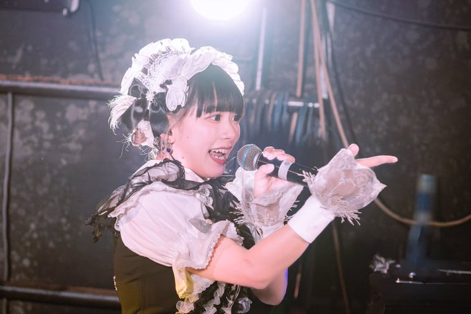 A female performer wearing a frilly white maid-inspired headpiece and outfit sings energetically into a microphone while pointing on a stage with bright lighting.