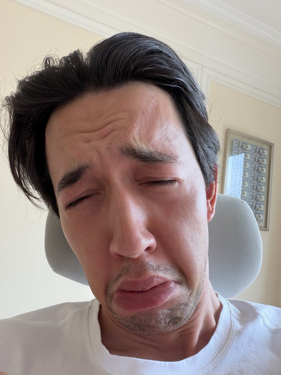Close-up selfie of a man with dark hair who appears to be dozing off or sleeping while wearing a gray neck pillow in front of a beige wall with a framed picture.