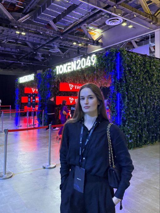 A young woman with long brown hair wearing a black shirt and lanyard poses in front of a 'TOKEN2049' entrance sign illuminated with neon lights and greenery at a tech conference.