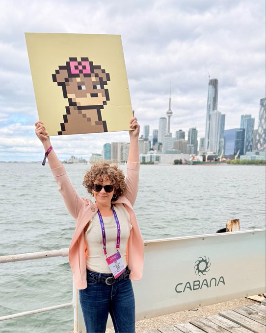 A curly-haired woman in a pink blazer holds up a pixel art painting of a bear character with a pink bow, photographed on a dock with the Toronto skyline visible in the background and 'CABANA' written on a nearby sign.
