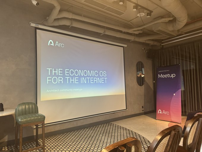 An indoor event space set up for an 'Architects | Meetup' hosted by Arc, with a projection screen displaying 'THE ECONOMIC OS FOR THE INTERNET' and an Arc-branded banner.