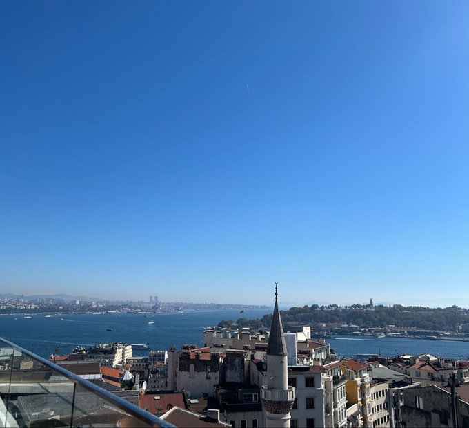 A scenic daytime view of a coastal city, likely Istanbul, showing the Bosphorus strait, boats, and a minaret rising above the rooftops under a clear blue sky.