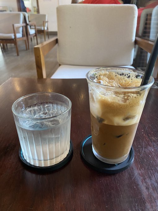 Two drinks on a wooden table in a cafe: a tall plastic cup of iced coffee with a black straw next to a small glass of water, both on black coasters.