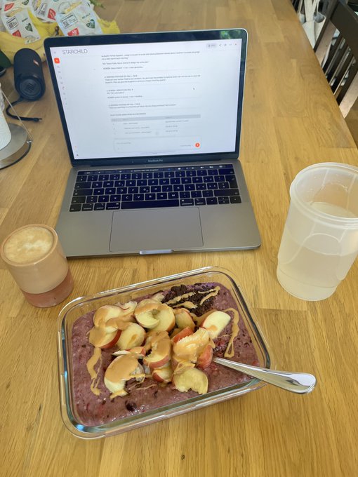 A glass dish of açaí topped with banana slices and peanut butter sits on a wooden table next to a MacBook Air displaying a Starlink webpage, with coffee and water nearby.