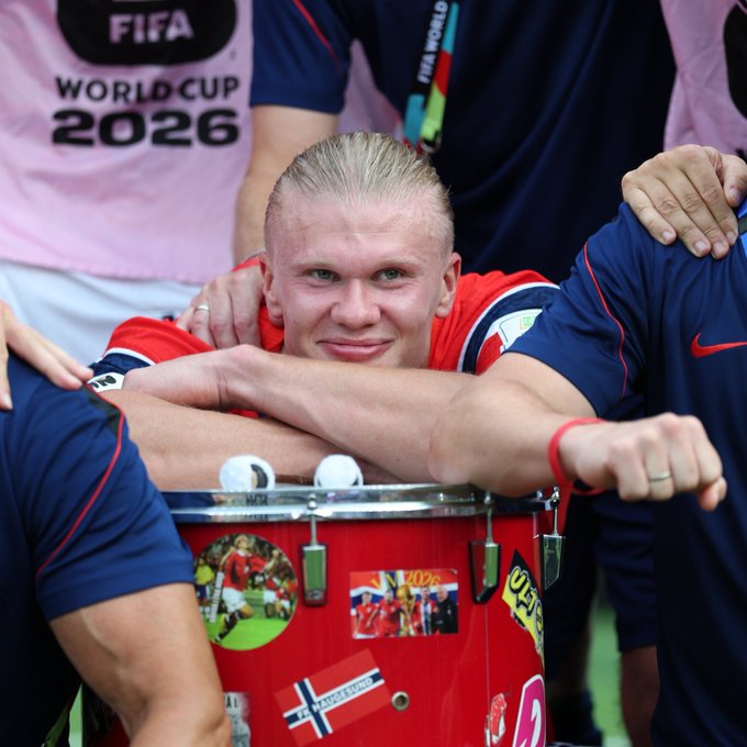 Erling Haaland smiles while leaning on a red drum decorated with flags, with teammates in pink FIFA World Cup 2026 shirts and navy blue jerseys around him.