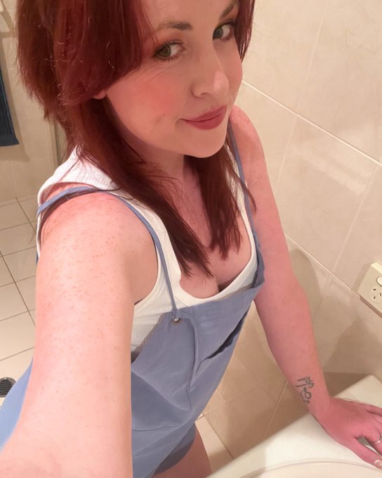 Selfie of a red-haired woman in a white tank top under blue overalls, sitting in a bathroom, showing freckles and a small tattoo on her wrist.