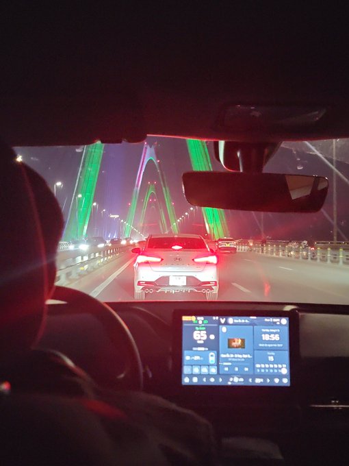 Nighttime photo taken from inside a car driving on a bridge illuminated with green lighting, showing the dashboard screen and a car ahead with red brake lights.