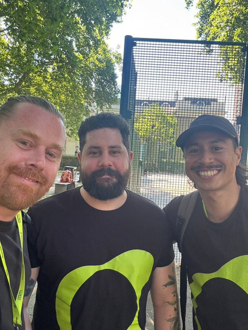 A selfie of three smiling men standing outdoors near a metal fence, with the two on the right wearing matching black t-shirts featuring a large lime green circular logo design.