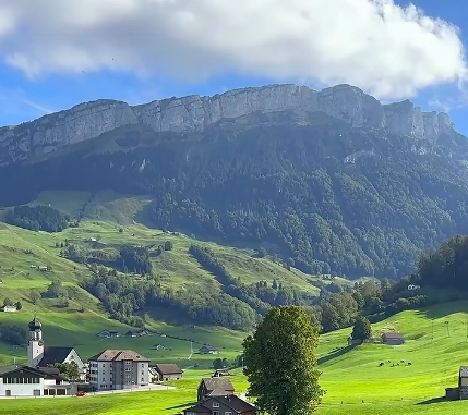 A scenic landscape view of a Swiss alpine village with green rolling hills, a church steeple, traditional houses, and a dramatic limestone cliff face in the background under a partly cloudy sky.