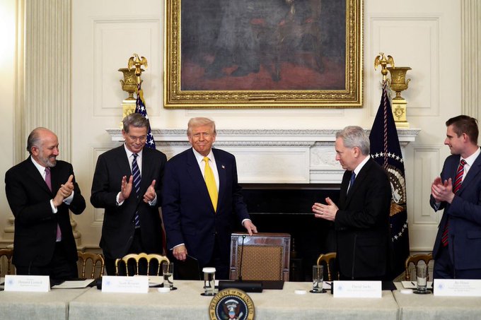 Donald Trump in a blue suit and yellow tie stands at a table in the White House Cabinet Room surrounded by applauding men in suits, with the Presidential seal on the table.