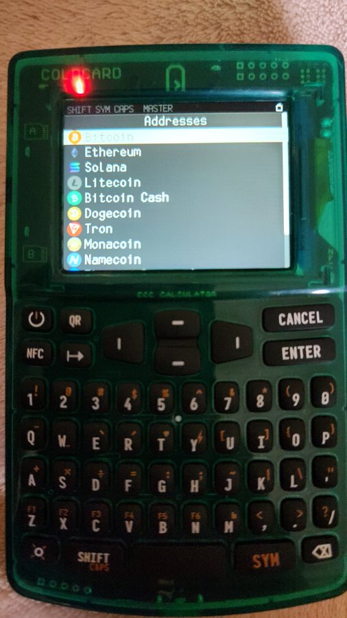 A close-up of a Coldcard hardware cryptocurrency wallet with a translucent green casing, displaying a menu of cryptocurrency addresses including Ethereum, Solana, Litecoin, and Bitcoin Cash on its screen.