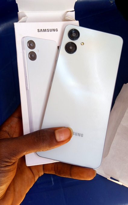 A hand holds a light blue Samsung smartphone in front of its open Samsung-branded box, displaying the dual rear camera setup and textured back design. The caption suggests a modest, aspirational purchase—possibly hinting at the buyer being a young person treating themselves to their first or new phone, with optimism that their situation will improve soon.