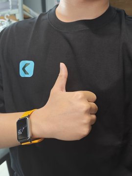 A person wearing a black t-shirt with a blue logo, giving a thumbs-up. They are wearing an Apple Watch with a yellow band displaying the time 9:41.