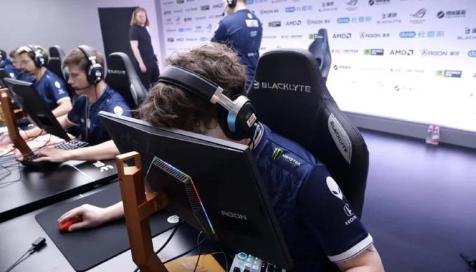 Esports players wearing headsets sit at gaming stations using monitors and peripherals, with 'AOC' and 'BLACKLYTE' branding visible on the equipment and 'Team Liquid' apparel on one of the players.