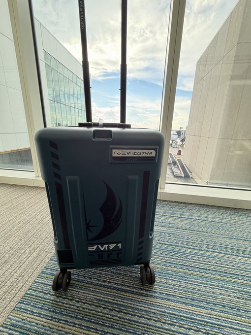 A dark teal suitcase branded with 'Mantic Games' logo and 'LIVE' text, sitting on a striped carpet in front of large windows showing an outdoor view.