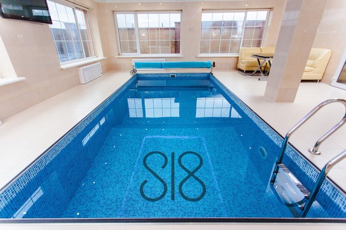 An indoor swimming pool with a monogram 'SJB' visible at the bottom, surrounded by tile flooring, windows, and a seating area.